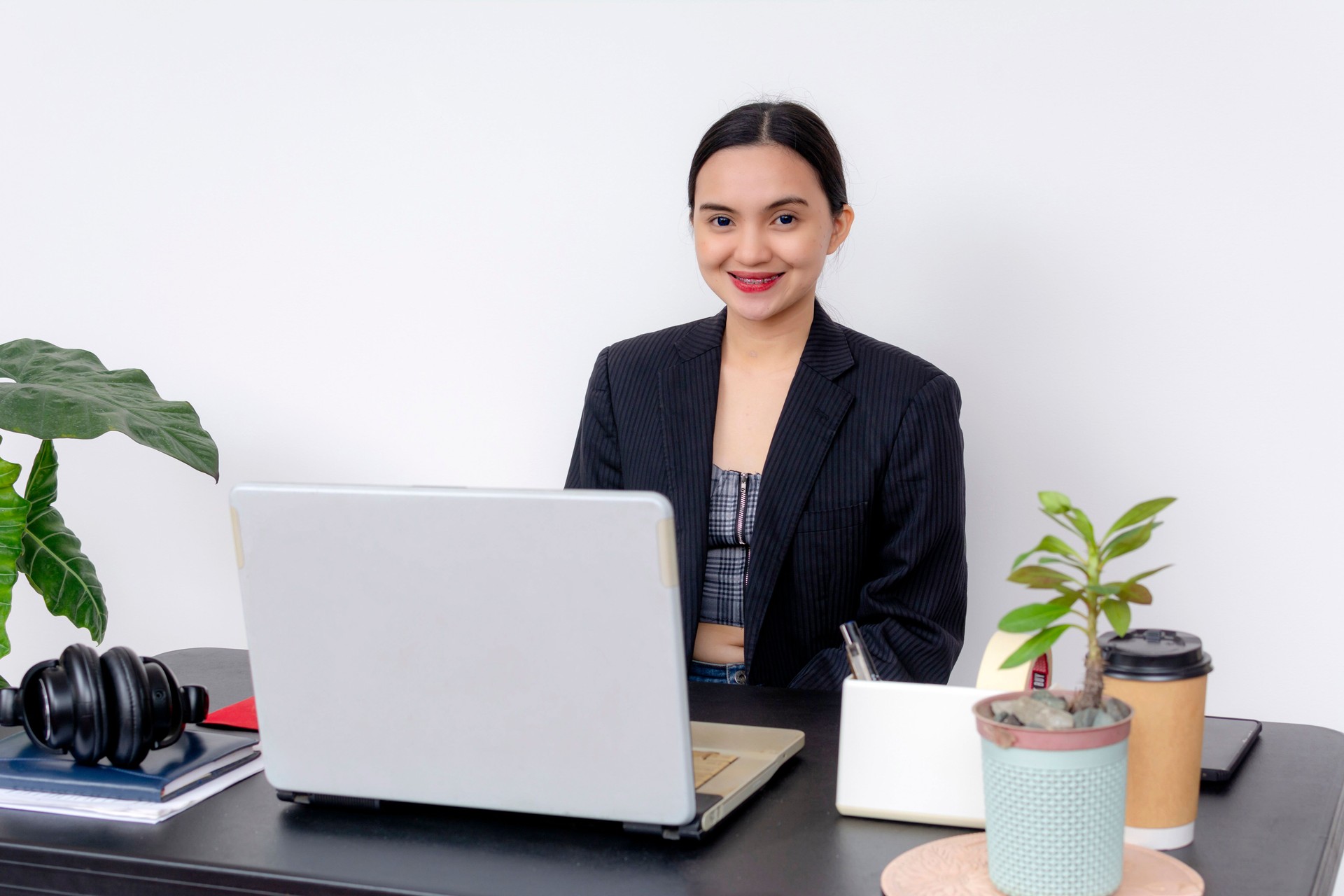 A young woman with a friendly smile works at her desk with a laptop, headphones, and coffee, projecting professionalism.